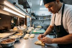 Um cozinheiro de avental e bandana preta trabalha concentrado na bancada de inox de uma cozinha profissional, moldando manualmente uma massa recheada (estilo esfiha ou pastel), cercado por bowls com ingredientes e porções de massa prontas.