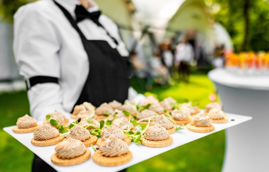 Garçom servindo uma bandeja com petiscos em um evento ao ar livre, com foco nos canapés decorados.