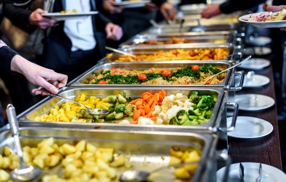 Um buffet com várias opções de comida, incluindo legumes, arroz e carnes, em um evento com pessoas servindo-se.