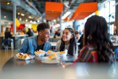 Grupo de jovens sorrindo e conversando em uma praça de alimentação colorida e movimentada enquanto comem lanches rápidos.