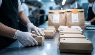 Profissional de luvas organiza embalagens de papel kraft em uma linha de montagem de delivery dentro de uma cozinha industrial movimentada.