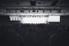 Grande plateia assistindo a uma apresentação ou palestra em um evento corporativo ou conferência, com iluminação focada no palco e telas de projeção ao fundo.