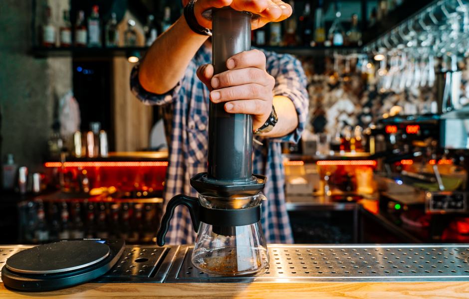 Um barista preparando café utilizando a técnica de infusão com a Aeropress em um ambiente de bar moderno. A imagem mostra a concentração e habilidade do profissional enquanto serve a bebida.