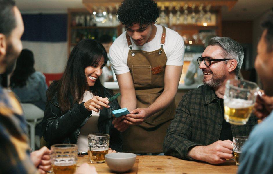 Atendente de avental auxilia clientes em um bar a realizar um pagamento ou pedido via dispositivo móvel.