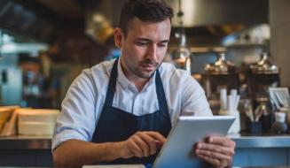 Homem de avental concentrado usando um tablet sobre um balcão de atendimento. O fundo mostra a cozinha de um restaurante ou café.