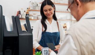 Jovem mulher sorridente fazendo pagamento com smartphone em uma cafeteria, usando uniforme e atendente ao lado, ambiente acolhedor e bem decorado