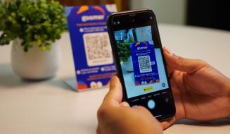 Pessoa escaneando um código QR com um celular na mesa, com uma embalagem azul ao fundo, promovendo a leitura de QR code para acesso a informações e ofertas.