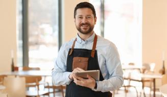 Homem sorridente usando um tablet em um ambiente de café, vestido com avental, mostrando a modernidade e a gestão de estabelecimentos comerciais.