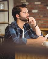Homem jovem de barba e camisa xadrez azul, sentado em uma mesa de café. Ele apoia a mão no queixo e olha para o lado com expressão séria. À sua frente estão um celular na mão, uma xícara de café e um tablet.