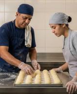 Profissionais de pastelaria/padaria trajando aventais, gorros de proteção e luvas transparentes organizam porções redondas de massa sobre tabuleiro metálico. Na bancada de inox sobressai farinha polvilhada, enquanto ao fundo observa-se parede revestida a azulejos claros com cartaz informativo.