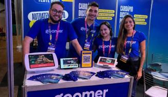 Quatro integrantes da equipe da Goomer, vestindo camisetas azuis com o logotipo da marca e crachás do evento, sorriem atrás de um balcão promocional. Sobre o balcão, há panfletos e diferentes dispositivos exibindo a plataforma de cardápio digital (totem, tablets e um display com QR Code).