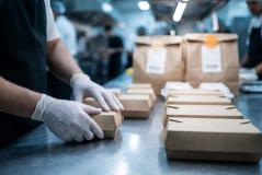 Profissional de luvas organiza embalagens de papel kraft em uma linha de montagem de delivery dentro de uma cozinha industrial movimentada.
