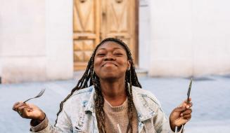 Mulher sorridente segurando garfo e faca ao ar livre, desfrutando uma refeição, com expressão de felicidade, em ambiente urbano. Uma cena que transmite alegria e satisfação ao comer.