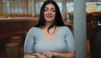 Mulher com cabelo preto e expressão de dúvida ou insegurança, vestindo camiseta azul clara, posando em ambiente de restaurante ou cafeteria.