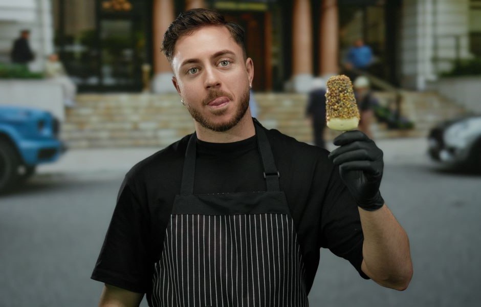 Jovem chef com barba e cabelo curto, vestindo avental preto listrado e luvas pretas, segurando um picolé de chocolate e limão em frente a um restaurante ou café ao ar livre.