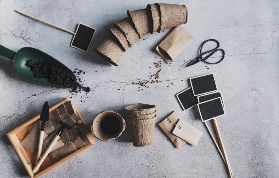 Materiais de jardinagem como vasos de papel, ferramentas e etiquetas estão organizados sobre uma superfície cinza, prontos para o cultivo de plantas.