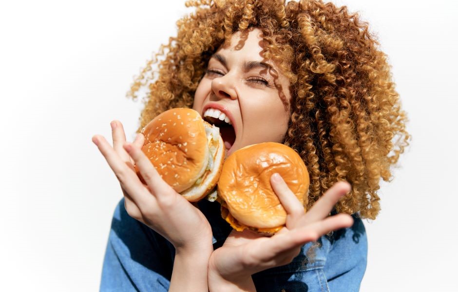 Mulher sorridente com cabelo cacheado dourado segurando dois hambúrgueres e expressando alegria,