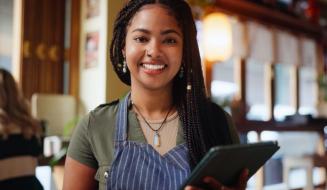 Mulher sorridente com cabelo trançado, usando avental no ambiente de um restaurante ou café, segurando um tablet, no contexto de trabalho na área de gastronomia ou atendimento ao cliente.