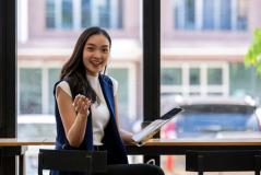 Mulher sorridente em um ambiente de trabalho moderno, sentada em uma mesa com um bloco de notas e um tablet, expressando entusiasmo e confiança.