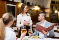 Jovem mulher atendendo clientes em um restaurante elegante, sorrindo e anotando pedidos, enquanto o homem na mesa olha para ela com interesse, com pratos e taças de vinho branco na mesa.