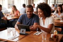 Casal está sentado em uma mesa de restaurante movimentado interagindo com um tablet fixado em um suporte. A mulher negra, de cabelo cacheado e camisa branca, aponta para a tela enquanto o homem ao seu lado observa atento. Sobre a mesa de madeira, há copos de água, talheres dispostos em guardanapos e um suporte com molhos.