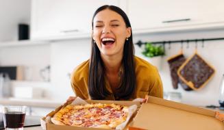 Mulher sorridente segurando uma caixa de pizza em uma cozinha moderna, transmitindo alegria e satisfação ao comer pizza.