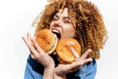 Mulher sorridente com cabelo cacheado dourado segurando dois hambúrgueres e expressando alegria,
