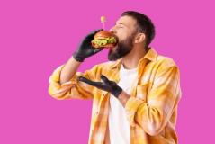 Homem jovem com barba e cabelo curto, usando jaqueta xadrez amarela e branco, segurando um hambúrguer com uma luva preta, pronto para comer, contra um fundo rosa vibrante.