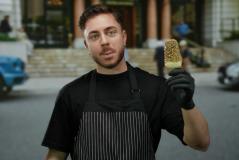 Jovem chef com barba e cabelo curto, vestindo avental preto listrado e luvas pretas, segurando um picolé de chocolate e limão em frente a um restaurante ou café ao ar livre.