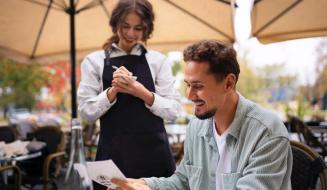 Garçonete anotando o pedido de um cliente sorridente que analisa o cardápio em um restaurante ao ar livre.