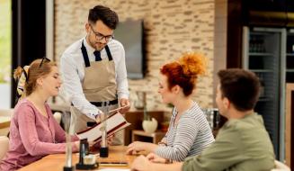 Garçom de avental bege e óculos atende três clientes sentados à mesa de um restaurante, apontando para as opções em um cardápio aberto.