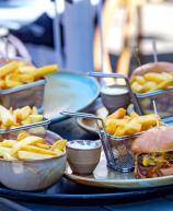 Imagem de uma refeição com batatas fritas e hambúrguer, incluindo molhos e acompanhamentos, servida em um ambiente ao ar livre.