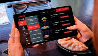 Pessoa usando um tablet para fazer pedido em um restaurante de comida japonesa, com pratos de sashimi e salmão visíveis na mesa, destacando o uso de tecnologia na experiência gastronômica.