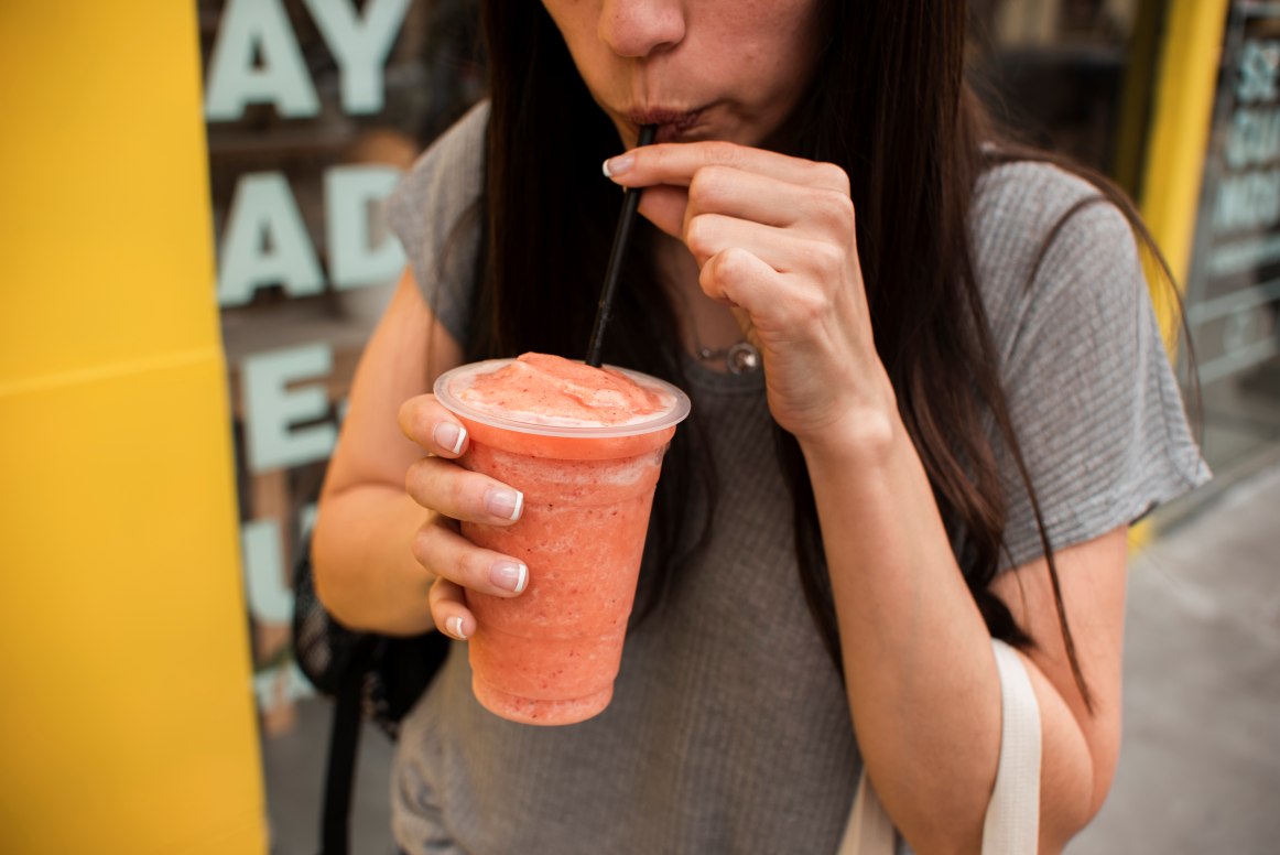 Close de uma mulher jovem saboreando um smoothie refrescante de frutas vermelhas (ou morango) através de um canudo. Uma imagem urbana e vibrante, perfeita para temas de estilo de vida saudável, refrescos de verão e bem-estar no dia a dia.
