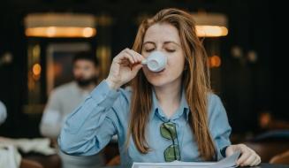 Mulher com cabelo castanho, usando blusa azul e óculos de grau presos à camisa, tomando uma xícara de café ou chá em um ambiente aconchegante, com foco na expressão de satisfação. Ao fundo, pessoas estão em um espaço moderno e bem iluminado, criando uma atmosfera de cafeteria ou sala de reunião, ideal para quem busca conforto e relaxamento.