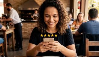 Cliente com cabelos cacheados sorri ao usar o celular para avaliar com cinco estrelas sua experiência no restaurante. Sentada à mesa com um prato de churrasco e uma bebida, ela aproveita o ambiente movimentado ao fundo, onde funcionários atendem outros clientes próximos à churrasqueira.