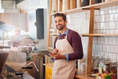 Homem sorridente com barba, vestindo avental e camisa azul, trabalhando em uma cafeteria ou café, segurando um tablet com prateleiras de produtos ao fundo.
