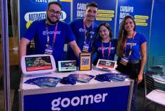Quatro integrantes da equipe da Goomer, vestindo camisetas azuis com o logotipo da marca e crachás do evento, sorriem atrás de um balcão promocional. Sobre o balcão, há panfletos e diferentes dispositivos exibindo a plataforma de cardápio digital (totem, tablets e um display com QR Code).