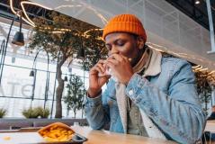 Homem com boné laranja saboreando um sanduíche em um café moderno, cercado por plantas e decoração contemporânea, com batatas fritas ao lado.