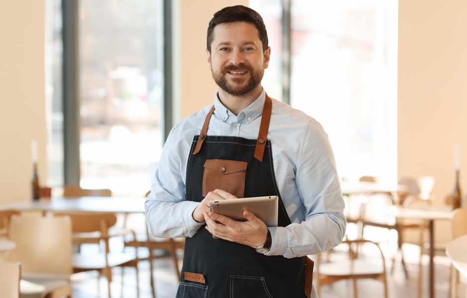 Homem sorridente usando um tablet em um ambiente de café, vestido com avental, mostrando a modernidade e a gestão de estabelecimentos comerciais.