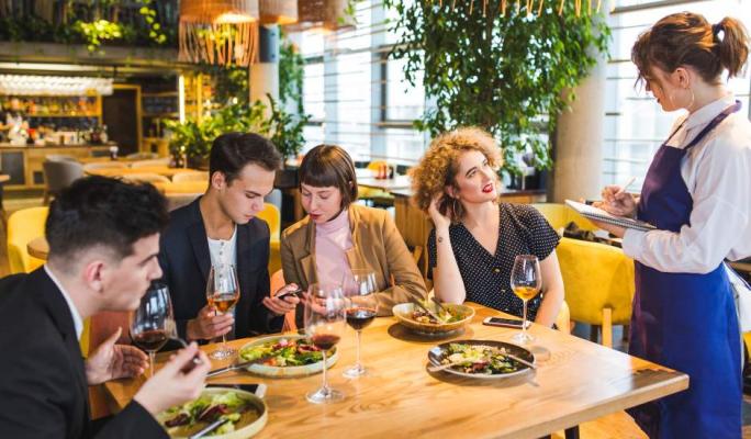 Garçonete de avental azul em pé anota o pedido em um bloco enquanto atende um grupo de quatro jovens sentados ao redor de uma mesa de restaurante com pratos de salada e taças de vinho.