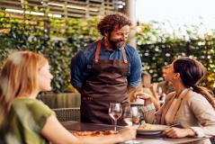 Homem com barba e cabelo cacheado atendendo duas mulheres em um restaurante ao ar livre, com pratos de comida e taças de vinho, ambiente acolhedor e natural.