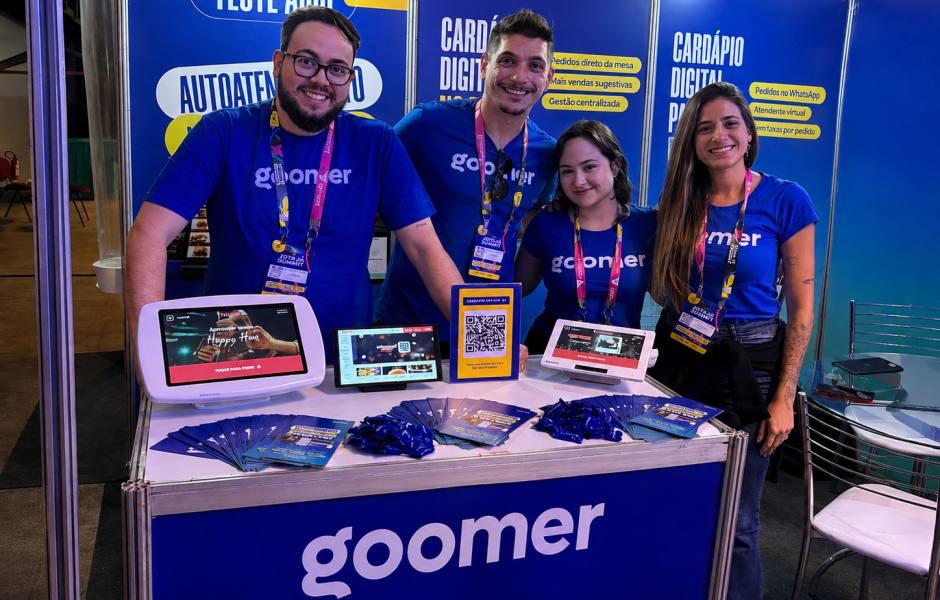 Quatro integrantes da equipe da Goomer, vestindo camisetas azuis com o logotipo da marca e crachás do evento, sorriem atrás de um balcão promocional. Sobre o balcão, há panfletos e diferentes dispositivos exibindo a plataforma de cardápio digital (totem, tablets e um display com QR Code).