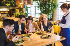 Garçonete de avental azul em pé anota o pedido em um bloco enquanto atende um grupo de quatro jovens sentados ao redor de uma mesa de restaurante com pratos de salada e taças de vinho.