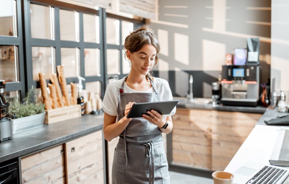 Jovem mulher atendente em cafeteria usando tablet, serviço de atendimento ao cliente, profissionalismo na cozinha com ambiente moderno e aconchegante.