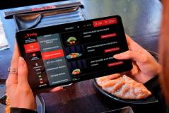 Pessoa usando um tablet para fazer pedido em um restaurante de comida japonesa, com pratos de sashimi e salmão visíveis na mesa, destacando o uso de tecnologia na experiência gastronômica.