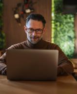 Homem de meia idade com barba, usando óculos, trabalhando em um laptop em um ambiente acolhedor e bem iluminado, com móveis de madeira e plantas ao fundo.