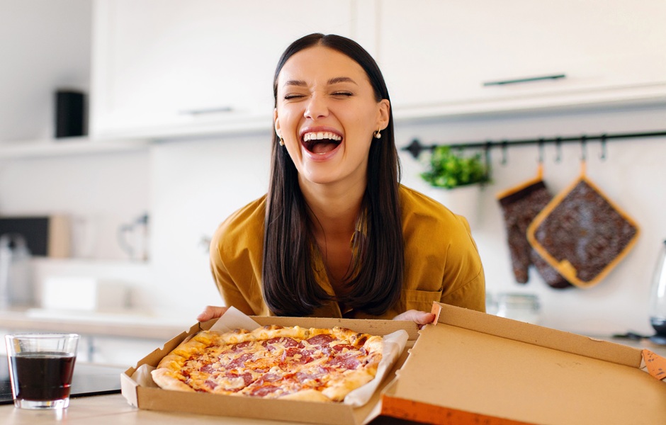 Mulher sorridente segurando uma caixa de pizza em uma cozinha moderna, transmitindo alegria e satisfação ao comer pizza.