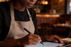 Mulher de avental escreve em tablet com caneta digital, simbolizando fazer a gestão do restaurante a distância do restaurante