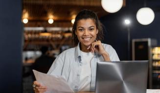 Mulher sorridente sentada à mesa com notebook e segurando papéis em um ambiente de escritório ou café.